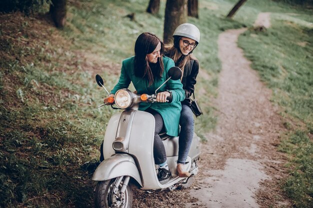 Un par de chicas hipster montando un scooter retro en la mujer hipster salvaje en una motocicleta italiana en el parque