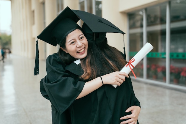 Par de alunas multi-asiáticas abraçando no dia da formatura no prédio da universidade se divertindo e dizendo adeus. conceito de diploma de vestido de qualificação educacional. amizade estilo de vida genuíno