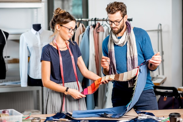 Foto par de diseñadores de moda eligiendo tela de pie en el estudio lleno de herramientas y equipos de sastrería