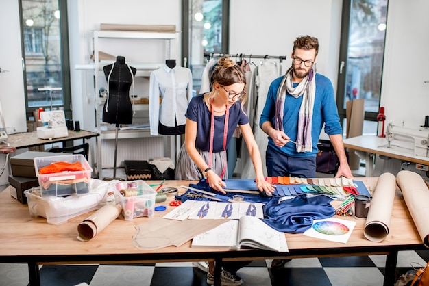 Par de diseñadores de moda que trabajan con bocetos de telas y ropa en el estudio lleno de herramientas y equipos de sastrería