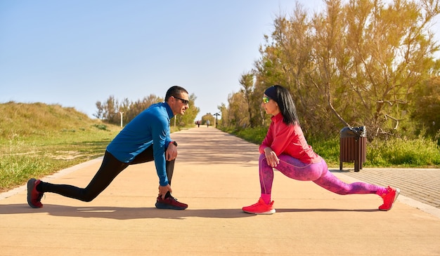 Par estirar las piernas antes de entrenar. La mujer viste ropas de color rosa brillante y violeta. El hombre lleva camisa azul y pantalón largo negro.