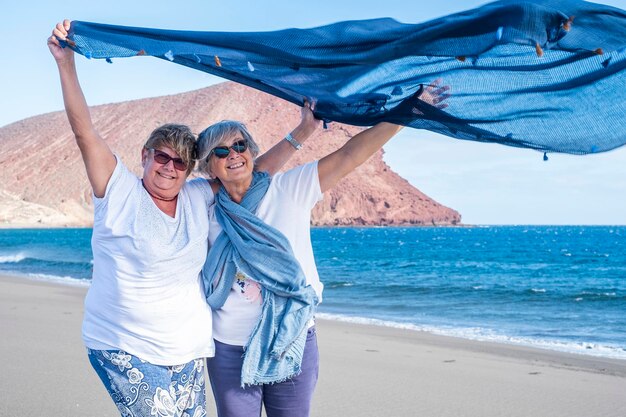 Un par de hermanas sonrientes y alegres disfrutando del mar de invierno en un día ventoso sosteniendo una bufanda en la brisa del mar. Personas mayores jubiladas activas