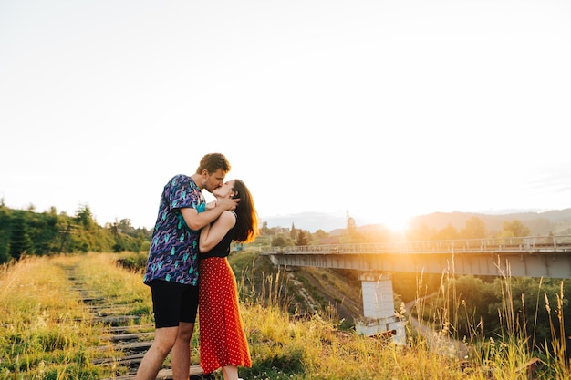 un par de jóvenes besándose de pie en un viaducto sobre un fondo de puesta de sol y un puente ferroviario