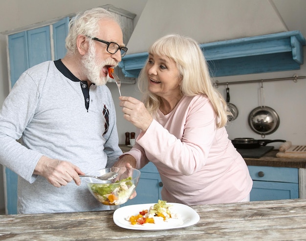 Un par de lindas personas mayores preparando comida. Linda anciana vestida de rosa claro alimentando a su marido con ensalada para el almuerzo.