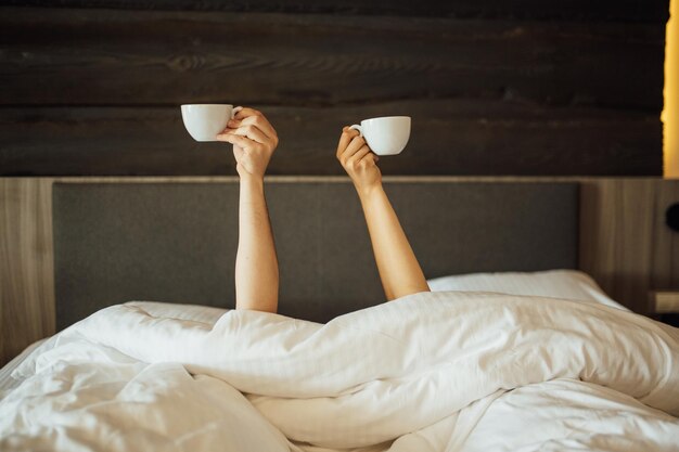 Un par de manos se sostienen sobre la manta en la cama y sostienen tazas de café. Tiempo de la mañana