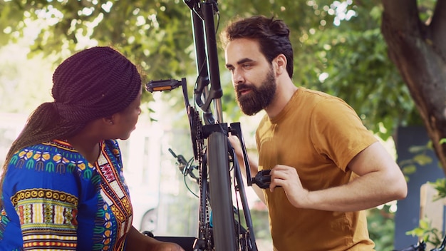 Par multirracial dedicado que fixa cuidadosamente a corrente e o pedal da bicicleta para ciclismo de lazer ao ar livre no verão. Casal jovem ao ar livre examinando minuciosamente os danos na roda da bicicleta para reparação.