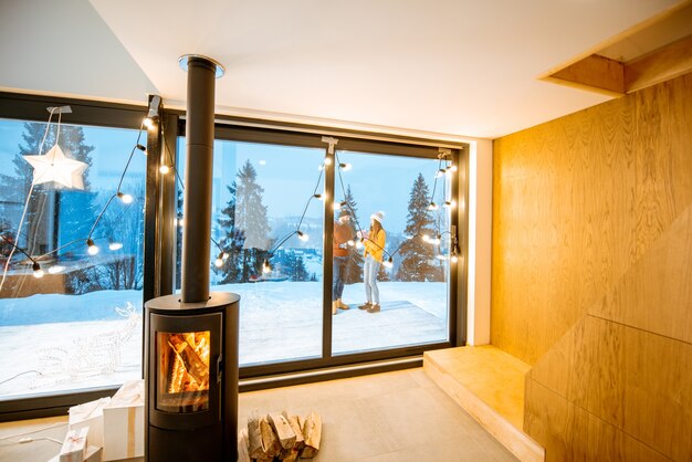 Par de pie al aire libre en la terraza de la casa con chimenea durante el invierno. Vista interior a través de la ventana.