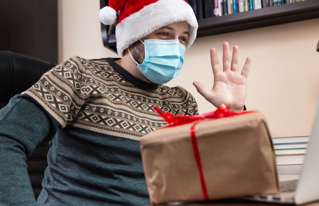 Foto parabéns online de natal. jovem com chapéu de papai noel e máscara facial dá um presente e fala usando o laptop para crianças e amigos de videochamada. natal durante o coronavírus.