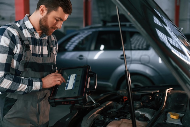 Foto parado perto do carro com o capô aberto segurando o tablet homem de uniforme está trabalhando no salão de automóveis