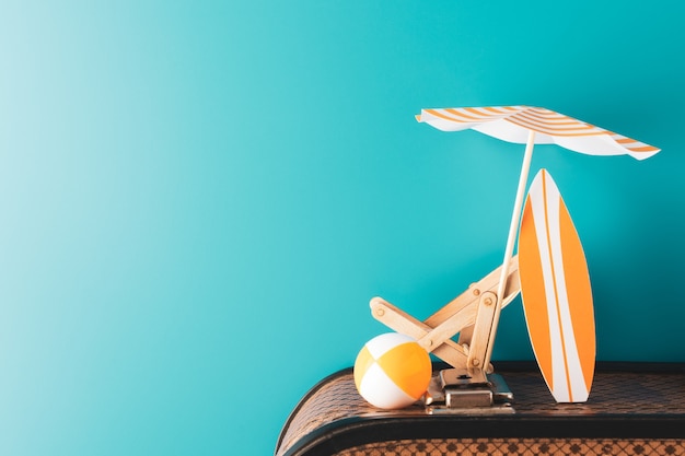 Paraguas de verano con chaise longue de madera, tabla de surf naranja y bola inflable en el equipaje sobre fondo azul.