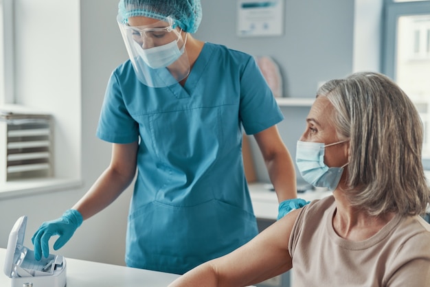 Paramédica feminina em trajes de trabalho de proteção, vacinando contra o coronavírus em mulheres maduras enquanto trabalhava no hospital