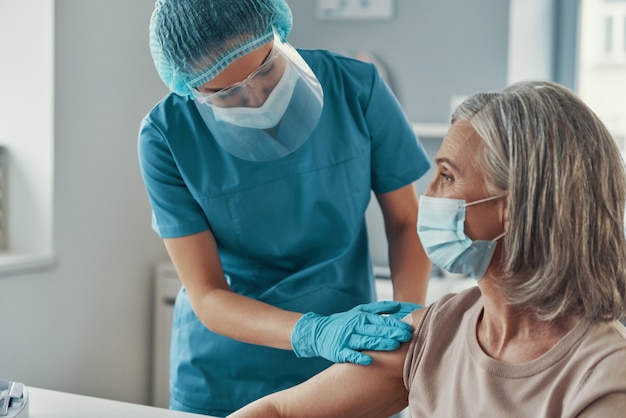 Paramédica feminina em trajes de trabalho de proteção, vacinando contra o coronavírus em mulheres maduras enquanto trabalhava no hospital