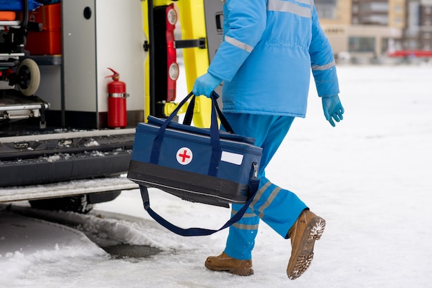 Paramédico masculino com roupa de trabalho azul e luvas carregando kit de primeiros socorros com cruz vermelha enquanto entra na ambulância