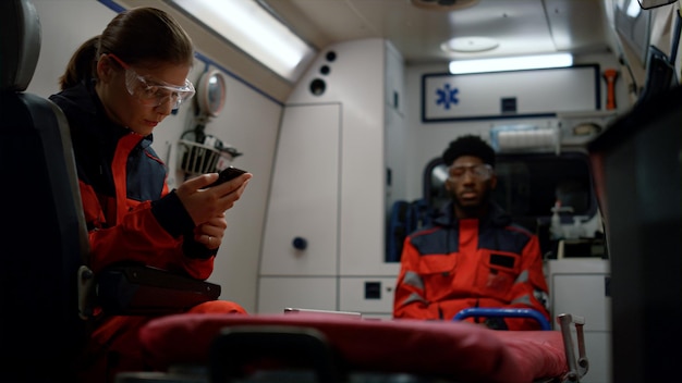 Foto paramedicos cansados sentados em um carro de emergência médica focada usando smartphone com óculos de proteção trabalhadores médicos multiétnicos conversando juntos ambulância conduzindo acidente de trânsito