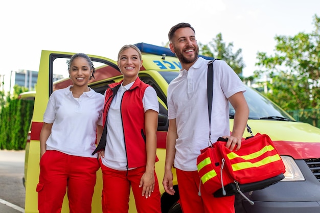 Foto paramédicos y médico de pie en la ambulancia lateral el médico lleva una bolsa de trauma médico grupo de tres paramédicos de pie frente a la ambulancia con una sonrisa