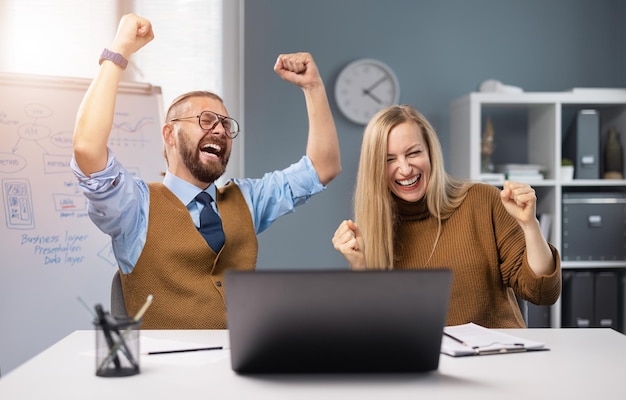 Parceiros de negócios caucasianos masculinos e femininos gesticulando emocionalmente de felicidade enquanto estão sentados na sala de escritório com laptop moderno sucesso e progresso no trabalho