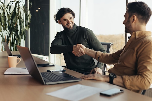 Parceiros de negócios felizes apertando as mãos de acordo fazendo um acordo sentado na mesa com o laptop no moderno