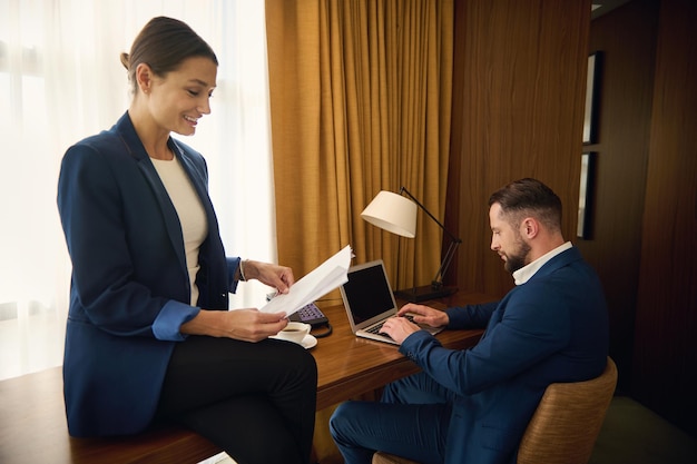 Parceiros de negócios trabalhando juntos em um quarto de hotel em viagem de negócios. Mulher de negócios, chefe com uma pilha de sorrisos de documentos em papel, contato de leitura, colega bonito sentado à mesa e trabalhando no laptop