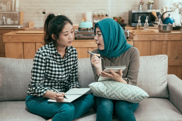parceiros de trabalho em equipe jovem asiática discutindo e fazendo anotações no sofá no campus da faculdade. estudantes muçulmanos e chineses estudando juntos sentados no sofá no dormitório. colegas de quarto do sexo feminino falando.