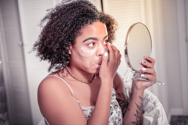 Parches debajo de los ojos. Mujer de pelo corto enfocada con lentes para los ojos mientras se mira en el espejo y tiene parches en la cara