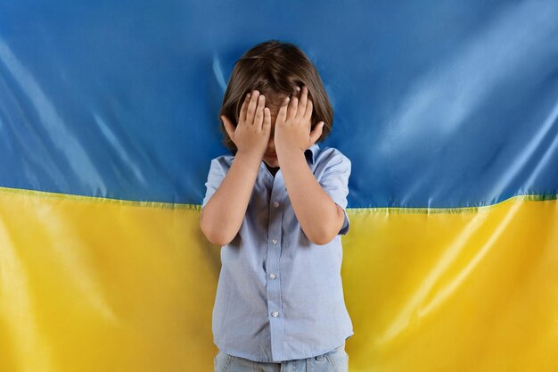 Pare de matar crianças na guerra Garotinho assustado chorando escondendo o rosto com as mãos posando sobre a bandeira ucraniana