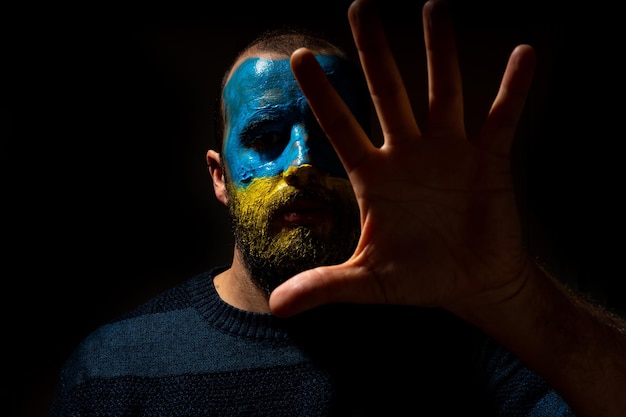 Pare o conflito de guerra entre a ucrânia e a rússia retrato de um jovem com o rosto pintado nas cores azul e amarela da bandeira fazendo o gesto para parar a guerra