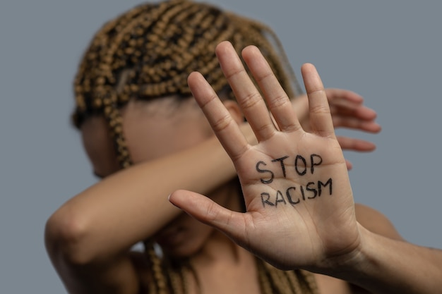 Foto pare o racismo. jovem afro-americana mostrando a palma da mão com a inscrição stop racism, cobrindo o rosto com o cotovelo