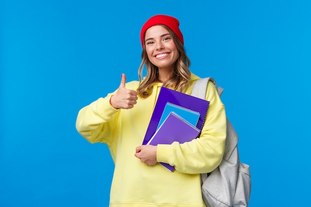 Parece bom Menina alegre bonita e inteligente caucasiana de gorro vermelho estudando muito para os exames, incentive o aluno a acompanhar, mostre o polegar para cima em aprovação ou gosto, segure o notebook e a mochila