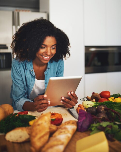 Parece que lo estoy haciendo todo bien Foto de una joven alegre navegando en una tableta digital mientras está rodeada de vegetales en la cocina de su casa durante el día