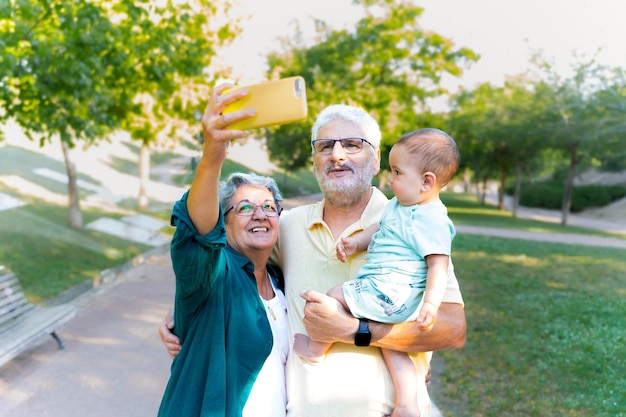 Pareja de abuelos felices con su nieto bebé en sus brazos tomando un autorretrato con su teléfono celular