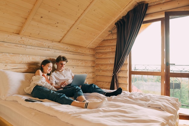 pareja acostada en la cama en un hotel con interior de madera y viendo películas en una laptop con cara feliz