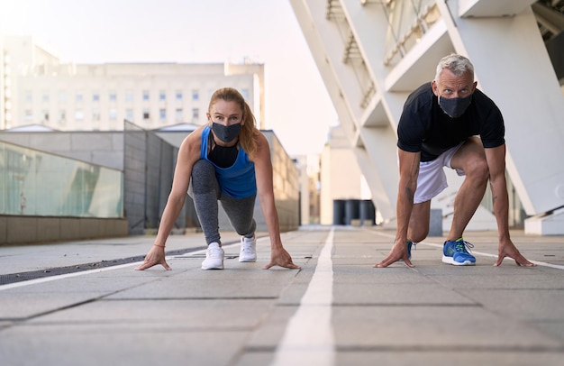 Pareja activa de mediana edad con máscaras protectoras en la posición de inicio lista para correr juntos