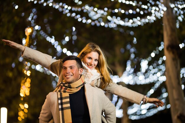 Pareja de adolescentes divirtiéndose Navidad Navidad por la noche al aire libre. Novio a cuestas novia con manos vuelan.