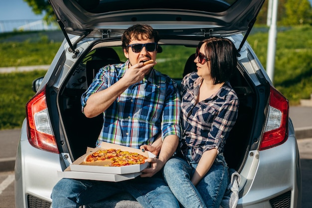 Pareja adulta elegante sentada en el maletero del coche y comiendo pizza al aire libre