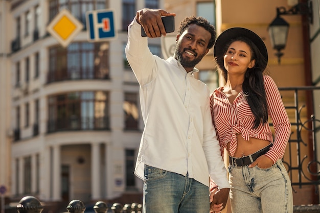 Pareja adulta joven tomados de la mano en la calle y sonriendo mientras toma selfie juntos