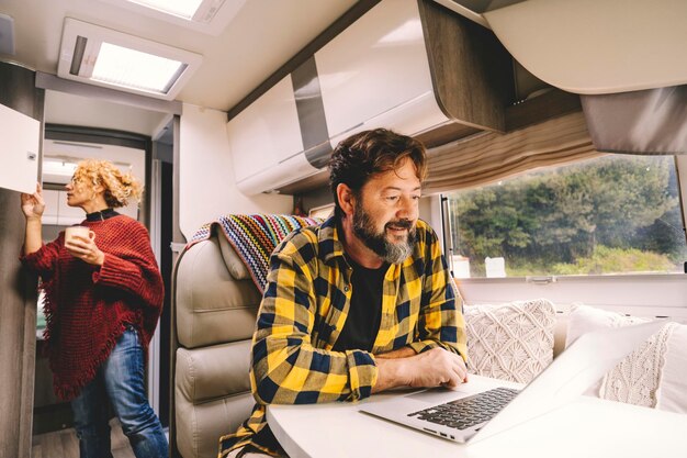 Una pareja adulta viaja y disfruta de una autocaravana para unas vacaciones de vacaciones o un estilo de vida de vida en una camioneta - hombre feliz trabaja en la computadora portátil sobre la mesa y mujer en busca de cosas dentro del vehículo - naturaleza al aire libre