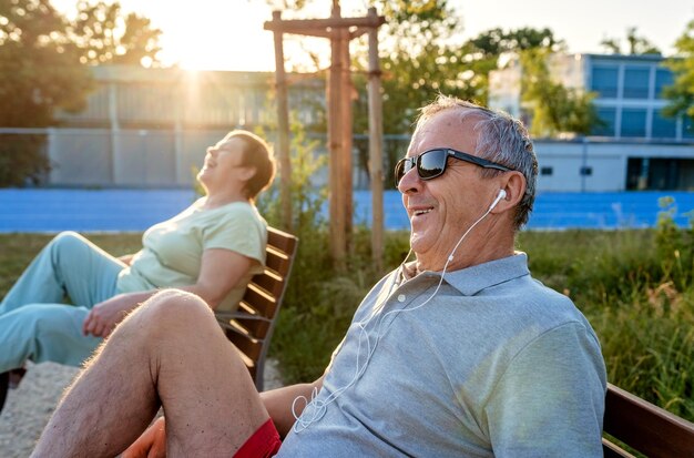 Foto una pareja de adultos mayores feliz y sonriente con auriculares haciendo ejercicio usando una máquina de bicicleta de entrenamiento al aire libre