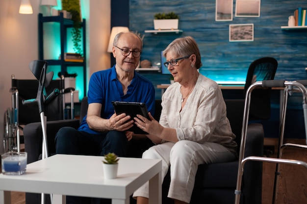 Pareja de adultos mayores con tableta digital en casa en el sofá con tecnología moderna para el entretenimiento y la comunicación por Internet en línea. Anciano y mujer con muletas y andador