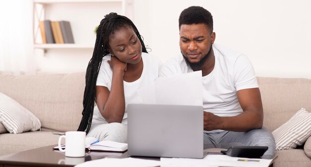 Pareja afro casada administrando juntos el presupuesto familiar en casa, leyendo documentos de hipotecas o seguros, panorama con espacio libre