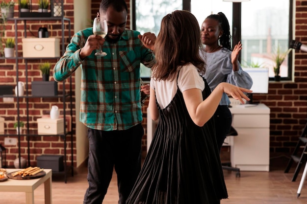 Pareja afroamericana bailando, disfrutando de la vida social acompañada de una joven caucásica adulta en una fiesta de amigos diversos. Gente de varias nacionalidades bebiendo vino, divirtiéndose.