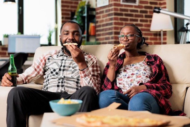 Pareja afroamericana comiendo pizza y bebiendo botellas de cerveza mientras ven películas en la televisión juntos. Disfrutando de comida rápida para llevar y bebidas, viendo televisión.