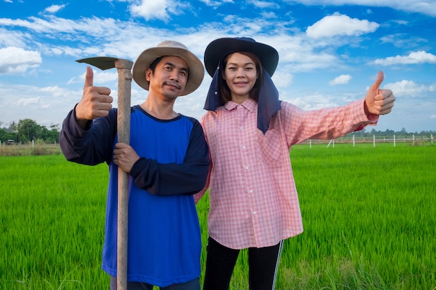 Pareja de agricultores asiáticos sosteniendo herramientas y pulgar arriba en granja de arroz