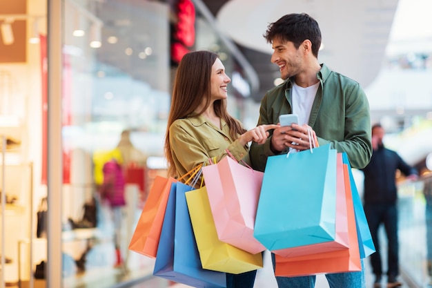 Una pareja alegre comprando por teléfono sosteniendo bolsas de compras en un hipermercado