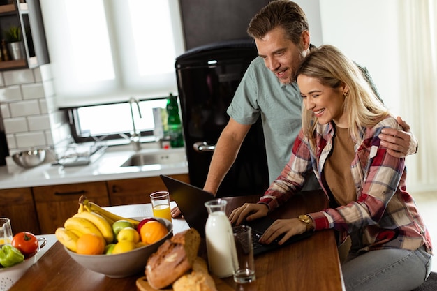 Una pareja alegre disfruta de un momento alegre en su cocina soleada trabajando en una computadora portátil rodeada de un desayuno saludable