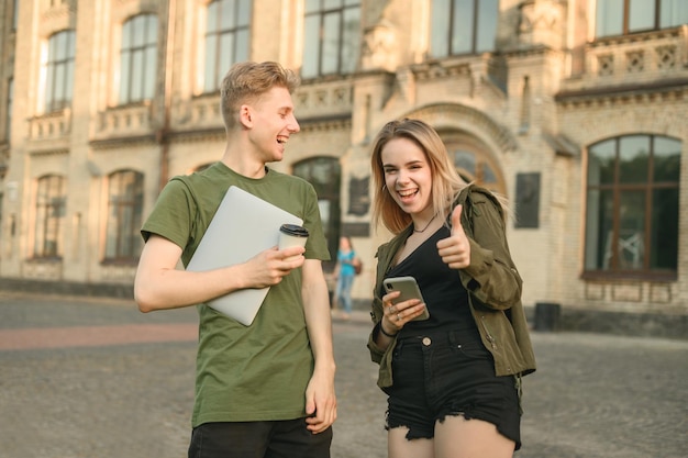 Una pareja alegre de jóvenes parados en la entrada de la universidad hablando y sonriendo sosteniendo una laptop