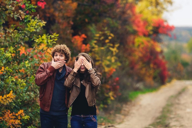 Pareja alegre muestra emociones hombre y mujer en chaquetas de cuero y jeans muestran sorpresa contra el fondo de los árboles de otoño