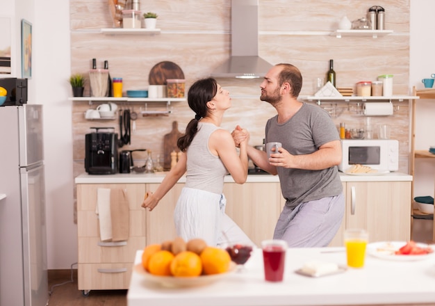 Pareja alegre en pijama bailando en la cocina escuchando música durante el desayuno. Marido y mujer despreocupados riendo, cantando, bailando escuchando meditando, viviendo felices y sin preocupaciones. Gente positiva. Han