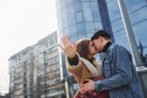 Una pareja alegre con ropa informal de abrigo besándose al aire libre en la ciudad cerca del edificio de negocios