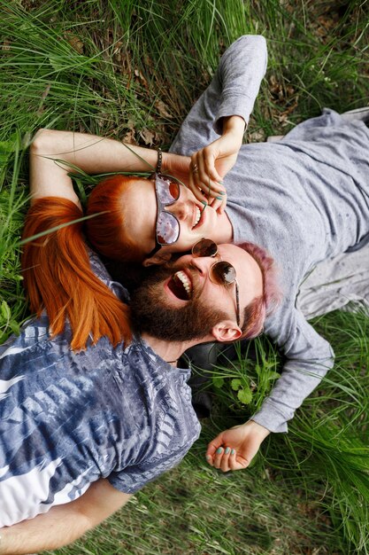 Pareja alegre, sentada sobre un césped verde, en el parque, sentada con la cabeza vuelta, con gafas de sol, mirando a la cámara.