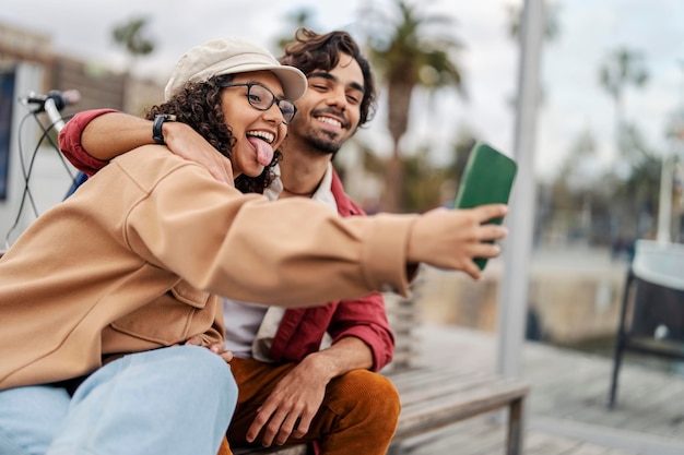 Una pareja alegre se toma selfies mientras se sienta en un banco en una calle de la ciudad